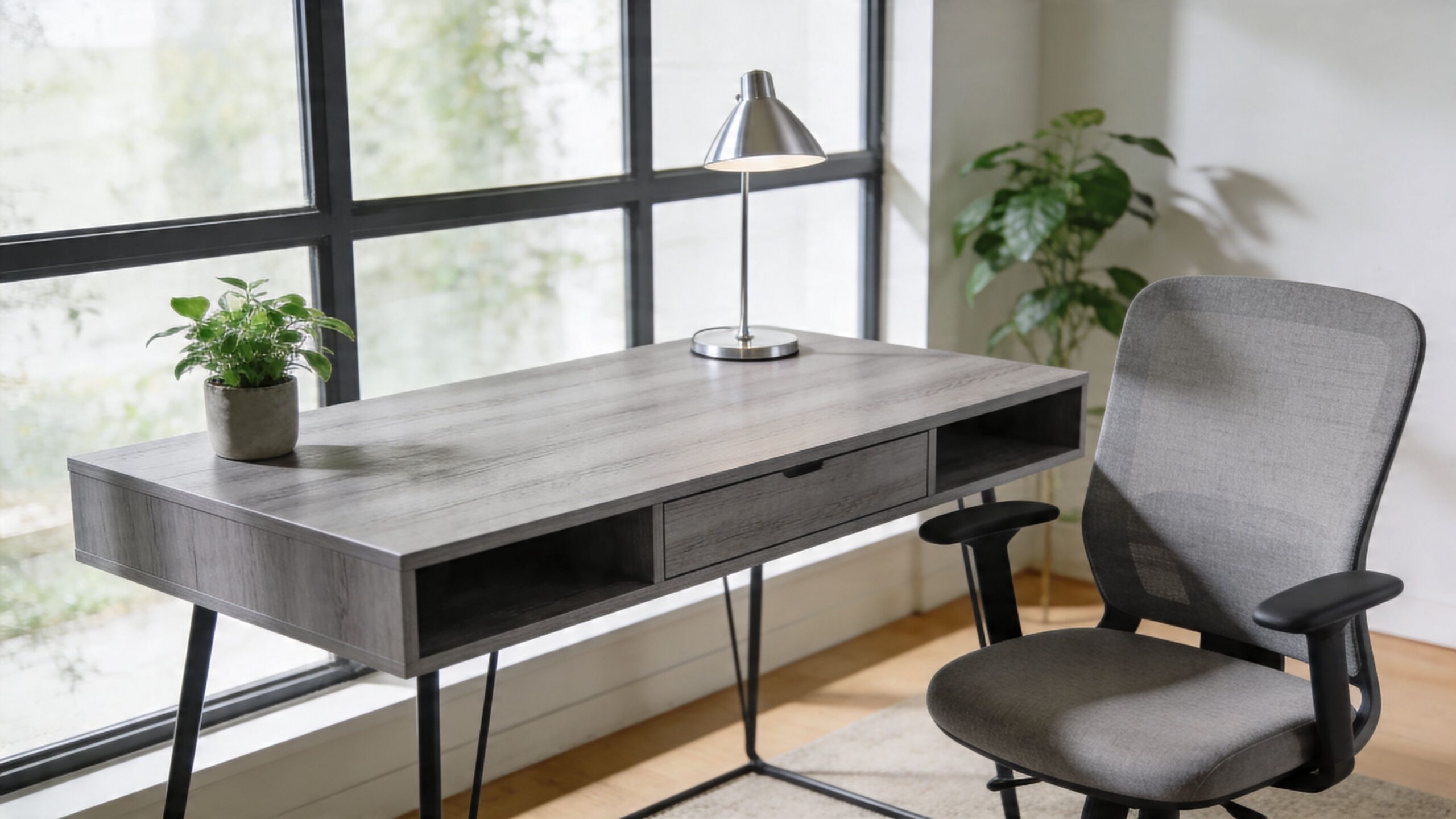 A modern grey wooden desk with a lamp, a small plant, and a grey office chair near a window.